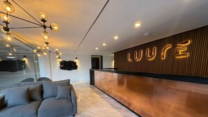 A modern reception area with a wooden reception desk, a grey sofa, and a chandelier. - HOTEL LUURE  (Huixquilucan)