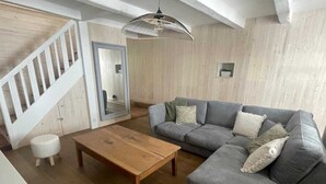 Una sala de estar con piso de madera, un sofá seccional gris, una mesa de centro de madera, un taburete blanco y una lámpara colgante.