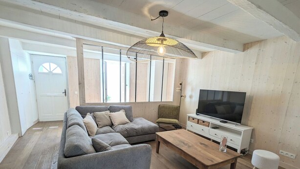Una sala de estar con un sofá seccional gris, una mesa de centro de madera, un televisor sobre un mueble blanco y una lámpara colgante.
