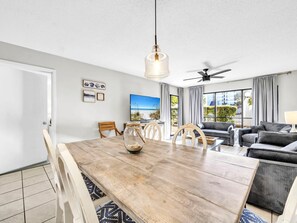 A modern living room with a wooden dining table, a ceiling fan, and a large window offering a view of the outdoors. - Edgewater Golf Villa 3204 (Panama City Beach)