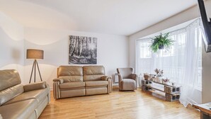 A living room with a beige leather sofa, a recliner, a floor lamp, and a large window with sheer curtains. - Le Cardinal Moderne-Wi-Fi rapide. Family-sized all included! (Laval)