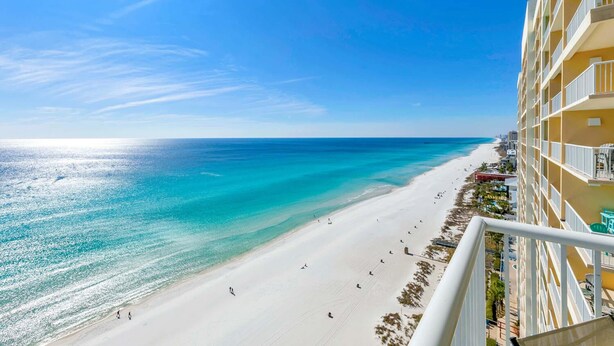 Blick auf einen Strand vom Hotelbalkon bei blauem Himmel, Sandstrand und wenigen Spaziergängern.