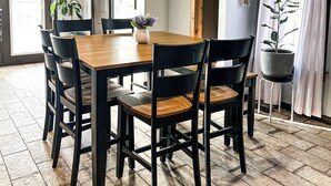 Un comedor con mesa de madera y sillas negras, una planta en maceta y vista al exterior a través de una ventana con cortinas.