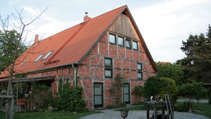 A two-story brick house with a red-tiled roof, a circular patio with a metal table and chairs, and a wooden fence. - The Village Island - Över de Deel (Trebel)