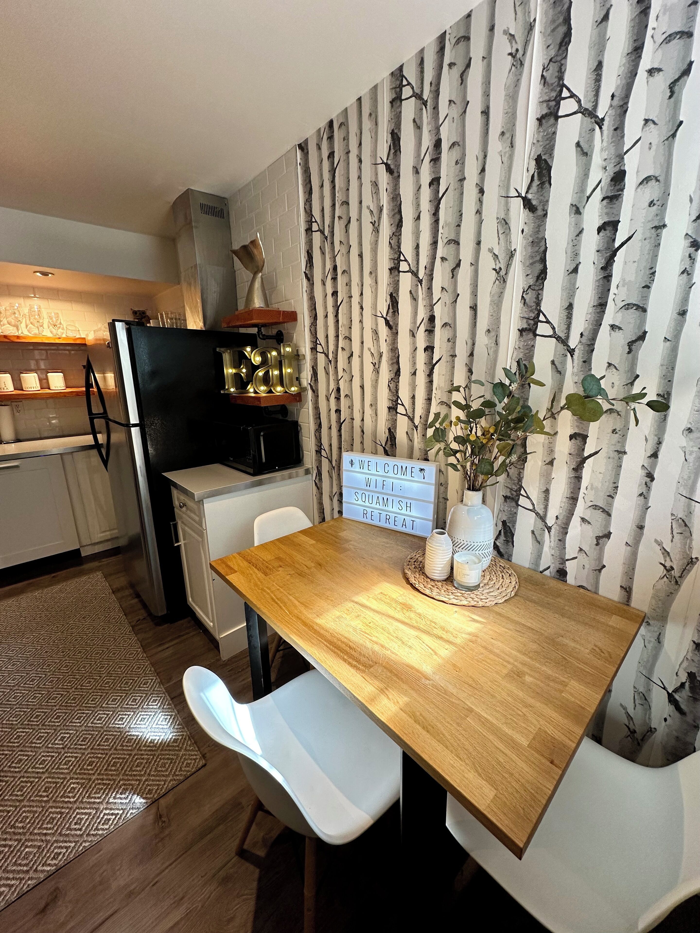 A modern kitchen with a refrigerator, stove, and white cabinets. There is a dining table with a placemat and a sign indicating WiFi and a squamish retreat.