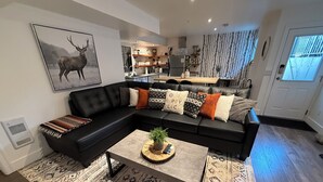 Ein modernes Wohnzimmer mit einer schwarzen Eckcouch, gemusterten Teppichen und einem zentral platzierten Couchtisch mit einer Pflanze.