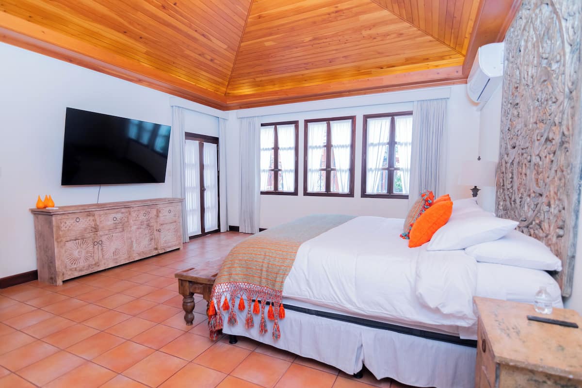 A bedroom with a large bed, a wooden ceiling, a TV mounted on the wall, and a stone wall.