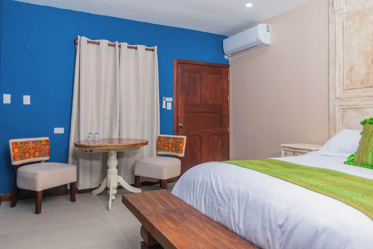 A hotel room with a bed, a wooden bench, a small round table with chairs, a curtain, and an air conditioning unit.