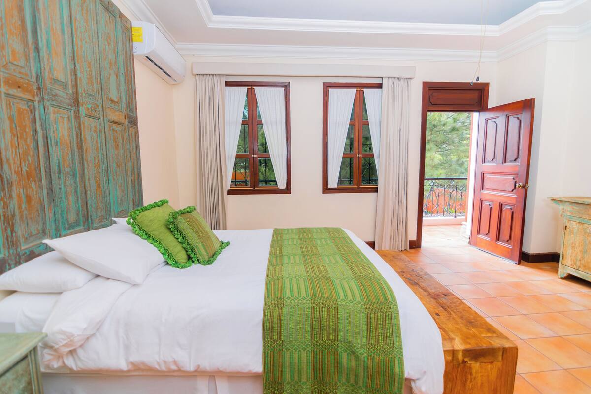A bedroom with a large bed, two windows with curtains, a wooden door, and a view of the outside.
