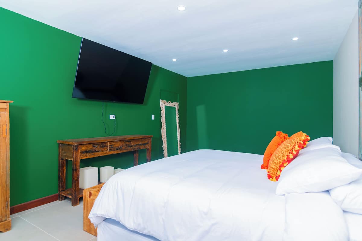 A room with green walls, a bed with white bedding and orange pillows, a wooden nightstand, a TV mounted on the wall, and a mirror.
