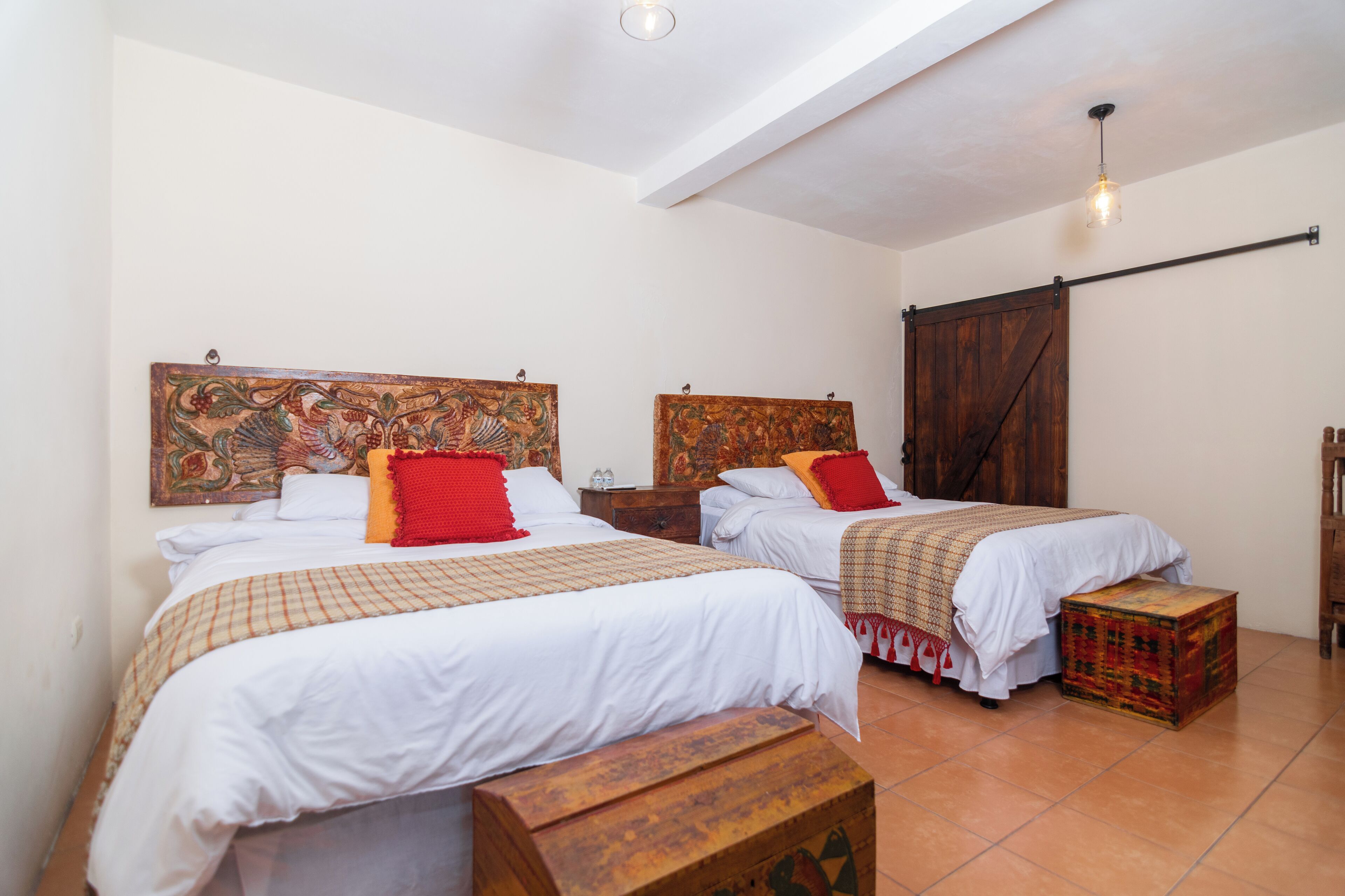 A room with two beds, each with a wooden headboard and white bedding. There is a wooden bench in the center and a small chest of drawers.