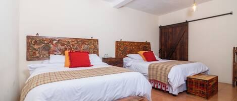 A room with two beds, each with a wooden headboard and white bedding. There is a wooden bench in the center and a small chest of drawers.