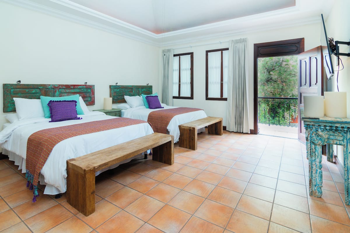 A hotel room with two beds, wooden benches, a balcony with a railing, and a table with a lamp.