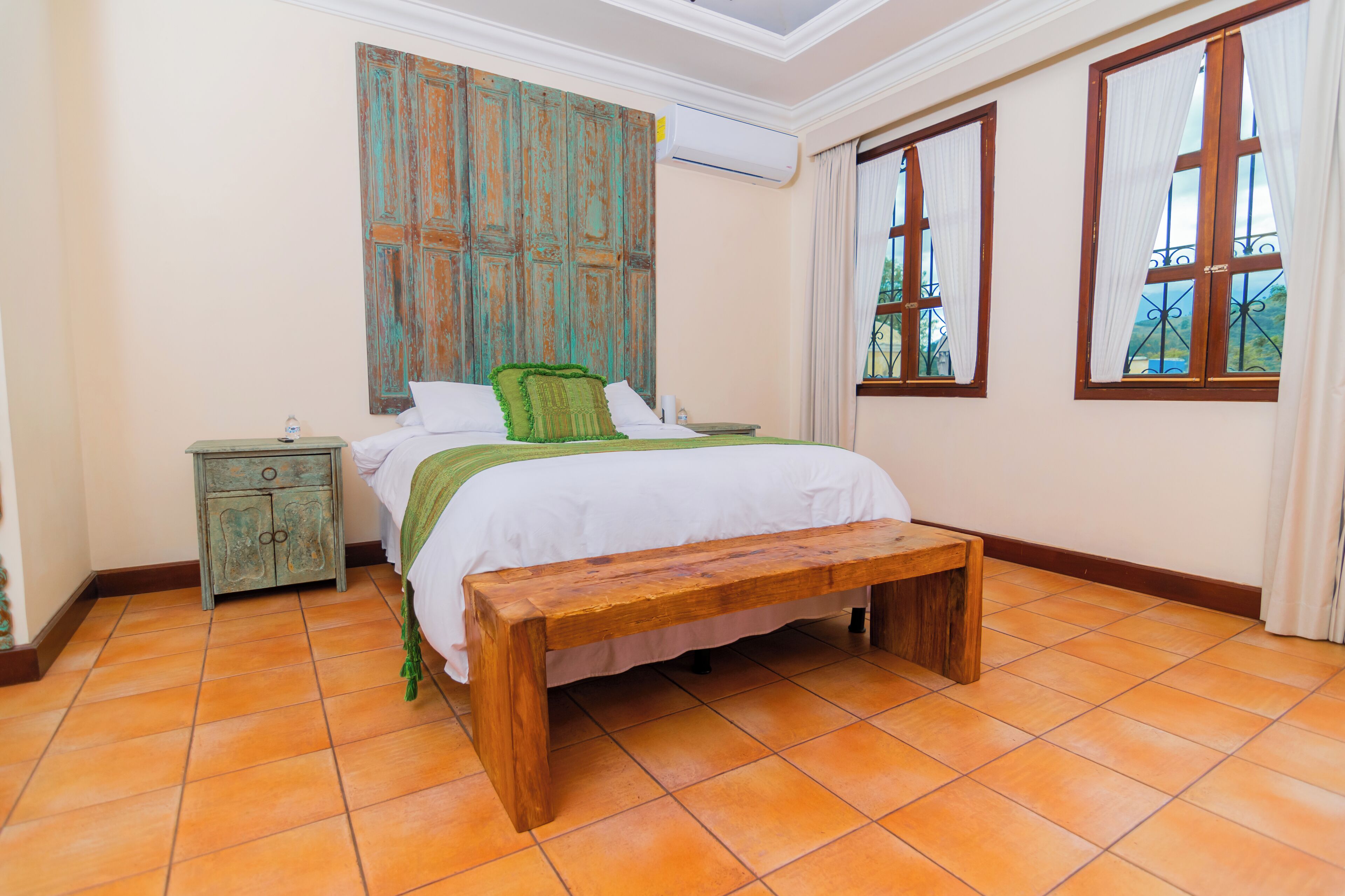 A bedroom with a wooden bed, a bench, a nightstand, and a large window with curtains.