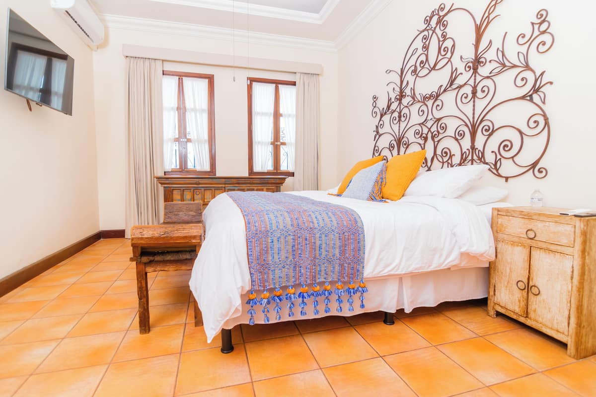 A bedroom with a bed, a wooden nightstand, a TV mounted on the wall, and a window with curtains.
