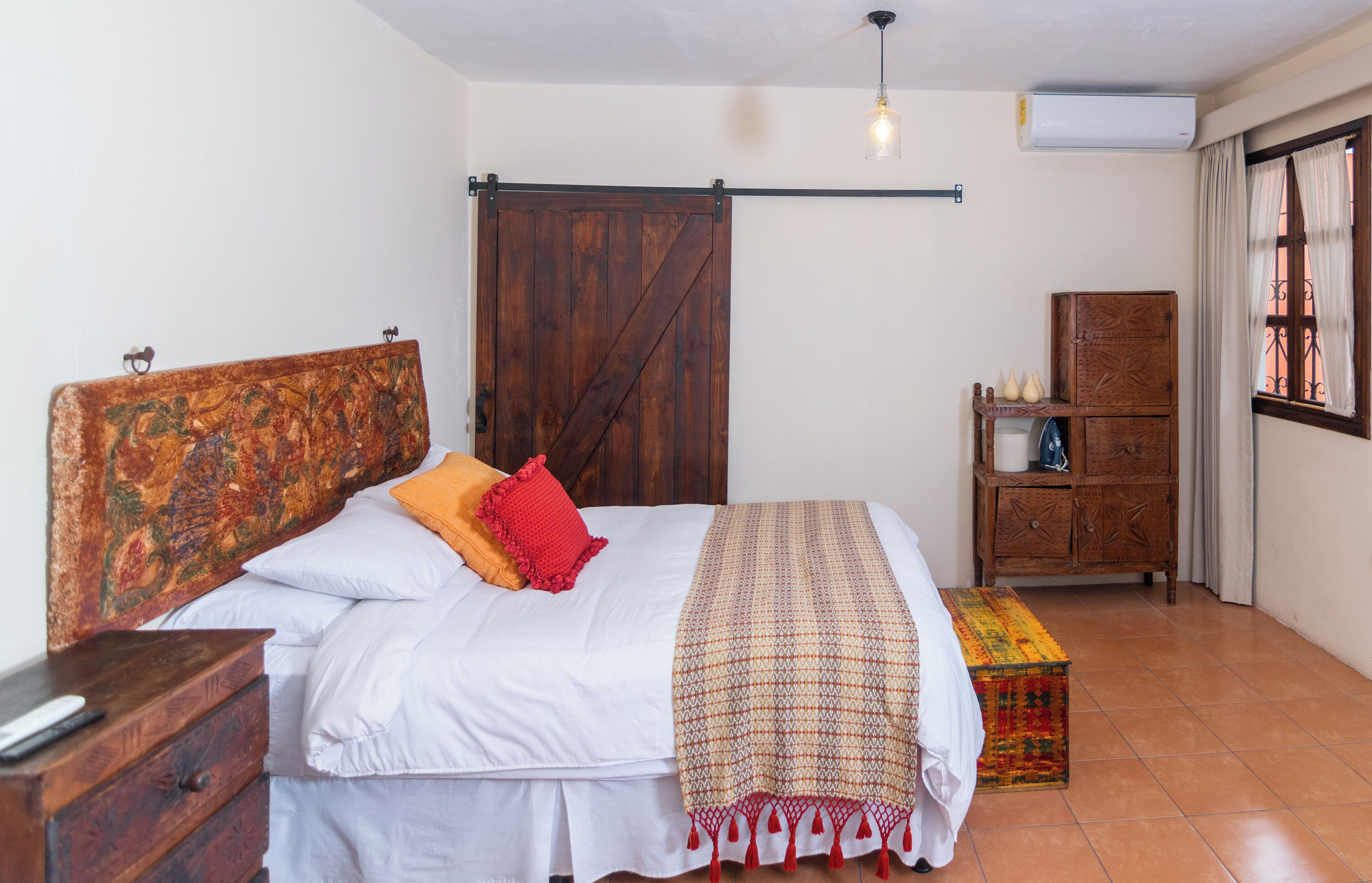 A bedroom with a wooden bed, a red pillow, a patterned blanket, a wooden chest of drawers, and a window with curtains.