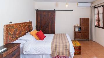 Een slaapkamer met een houten bed, een rood kussen, een bedekker met patroon, een houten dressoir en een raam met gordijnen.