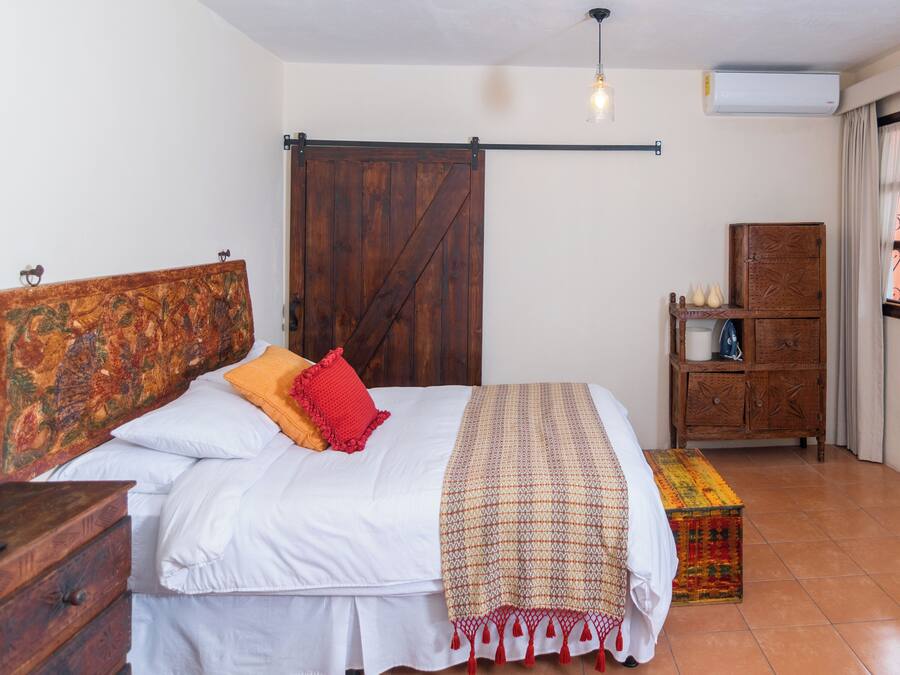 Een slaapkamer met een houten bed, een rood kussen, een bedekker met patroon, een houten dressoir en een raam met gordijnen.