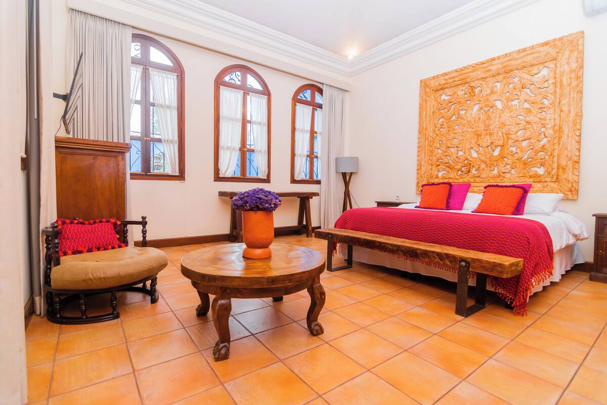 A room with a bed, a wooden bench, a round table with a vase, and two chairs.