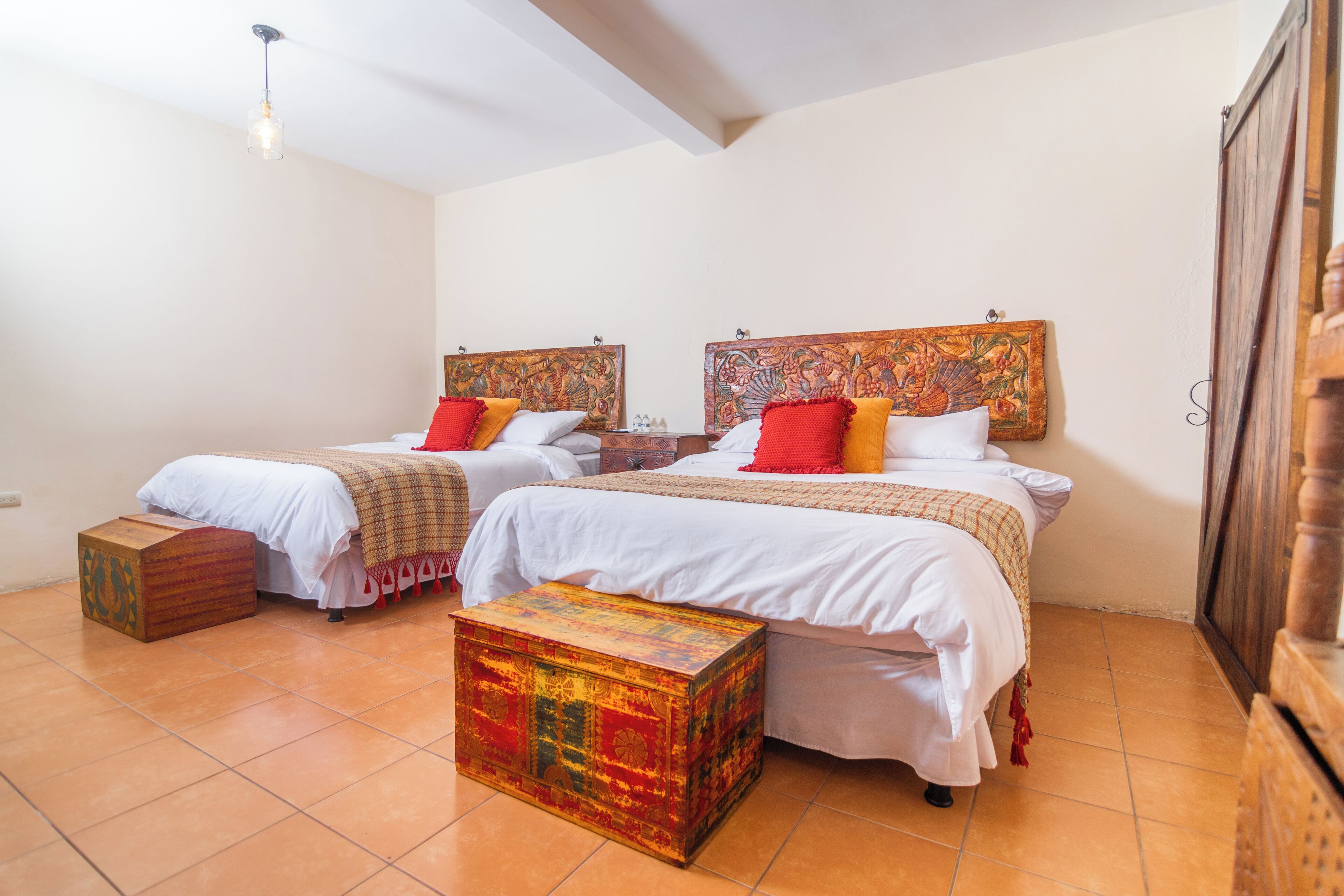 A room with two beds, each with a headboard, white bedding, and red and yellow pillows. There is a colorful chest of drawers and a small wooden stool.
