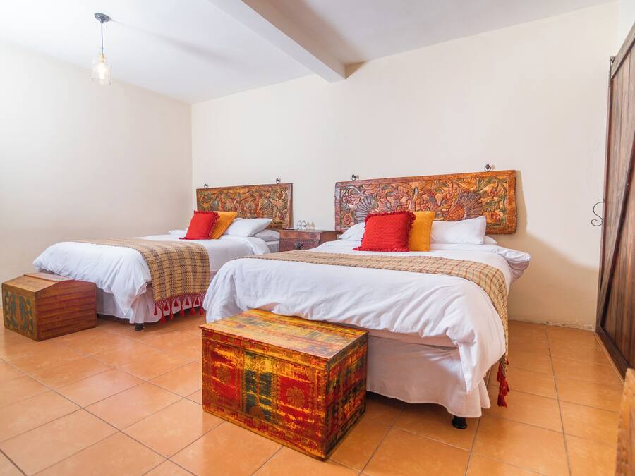 A room with two beds, each with a headboard, white bedding, and red and yellow pillows. There is a colorful chest of drawers and a small wooden stool.