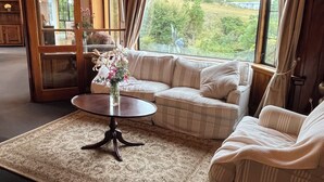 A cozy living room with a sofa, a coffee table, and a view of a green landscape through large windows. - Greenwood Homestead (Tarara)