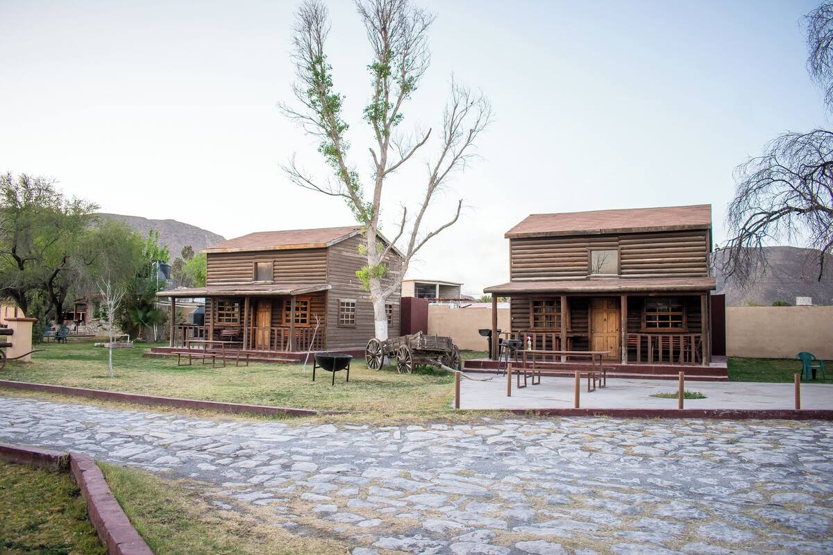 Edifici de fusta d'estil cabana rústica amb un pati empedrat de pedra, bancs de fusta i un carretó de fusta.