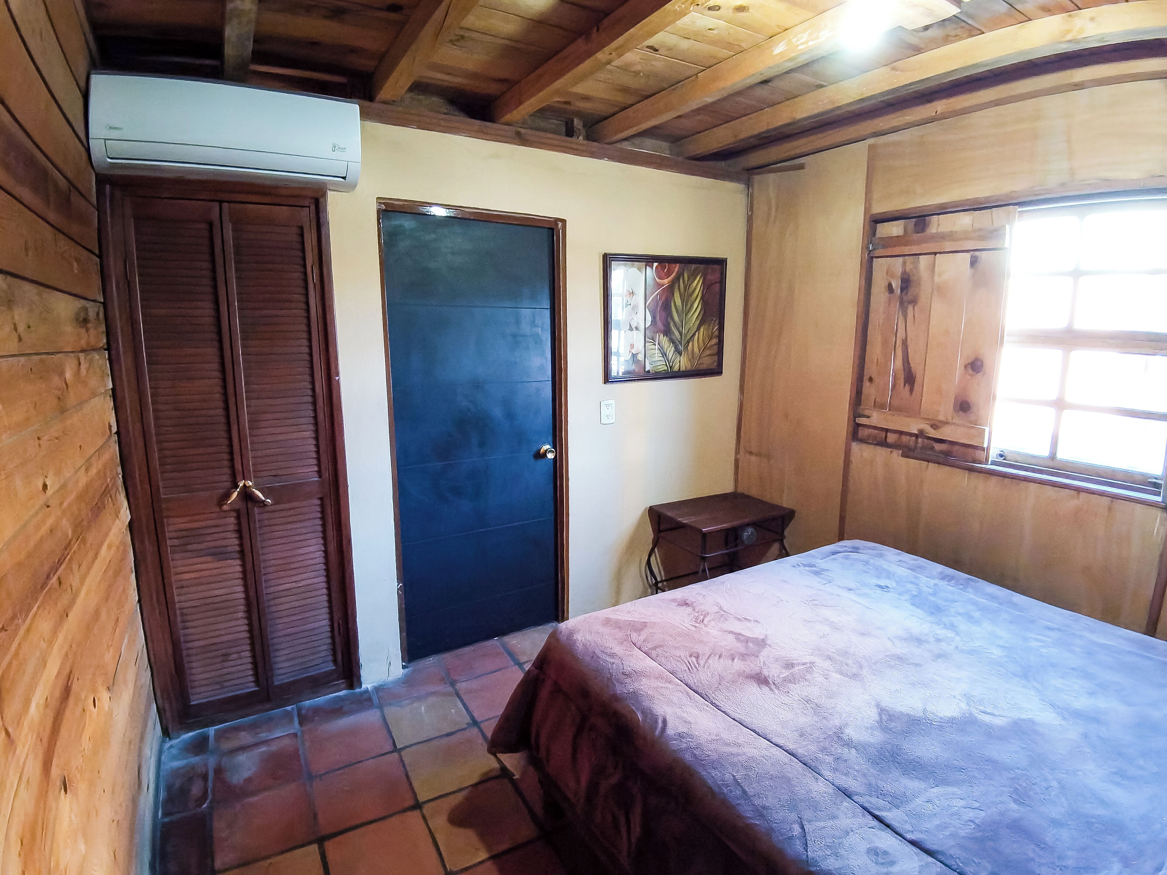A room with wooden walls and ceiling, a bed with a blanket, a door, a small table, and a window.