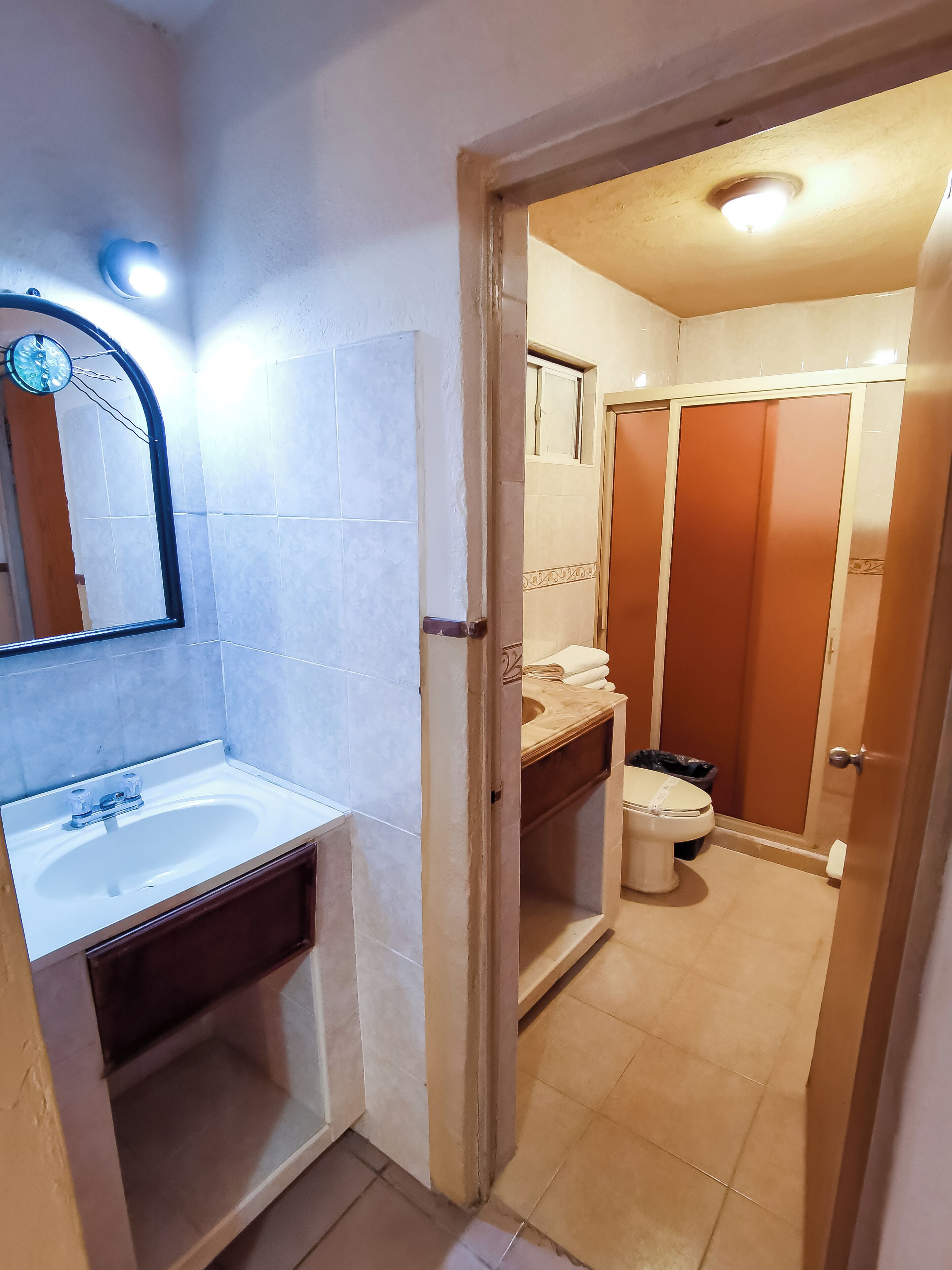 Ein Badezimmer mit einem weißen Waschbecken, Spiegel und gefliesten Wänden. Eine Holztür führt zu einem Duschbereich mit Toilette und einem Mülleimer.