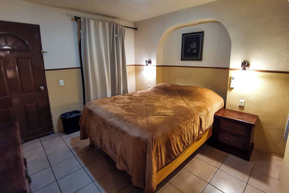 Un dormitori amb un llit, una tauleta de nit, una porta, una cortina i una làmpada penjada a la paret.