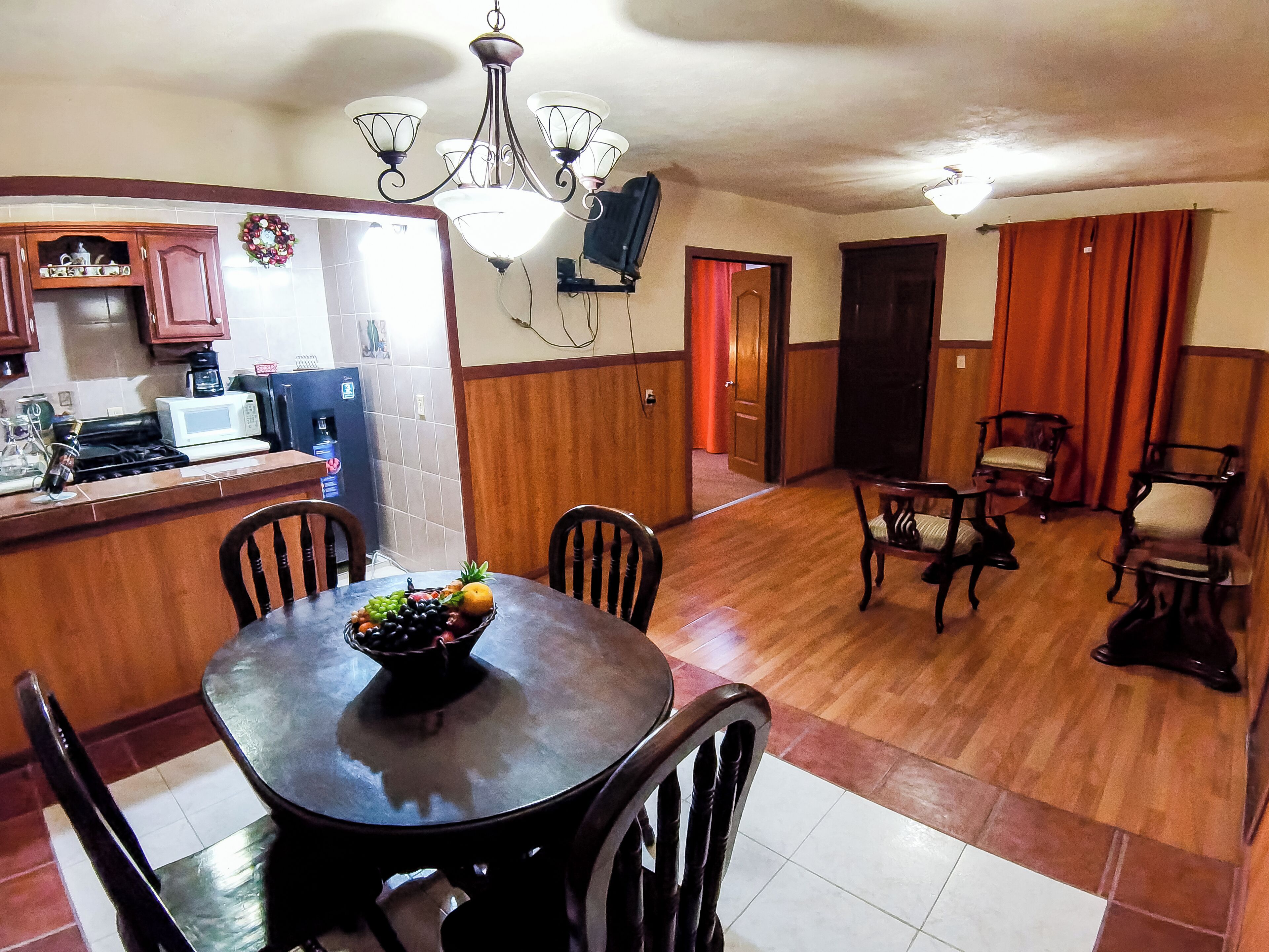 Un comedor con una mesa de madera, sillas y un frutero. Se ve una cocina al fondo con microondas y estufa.