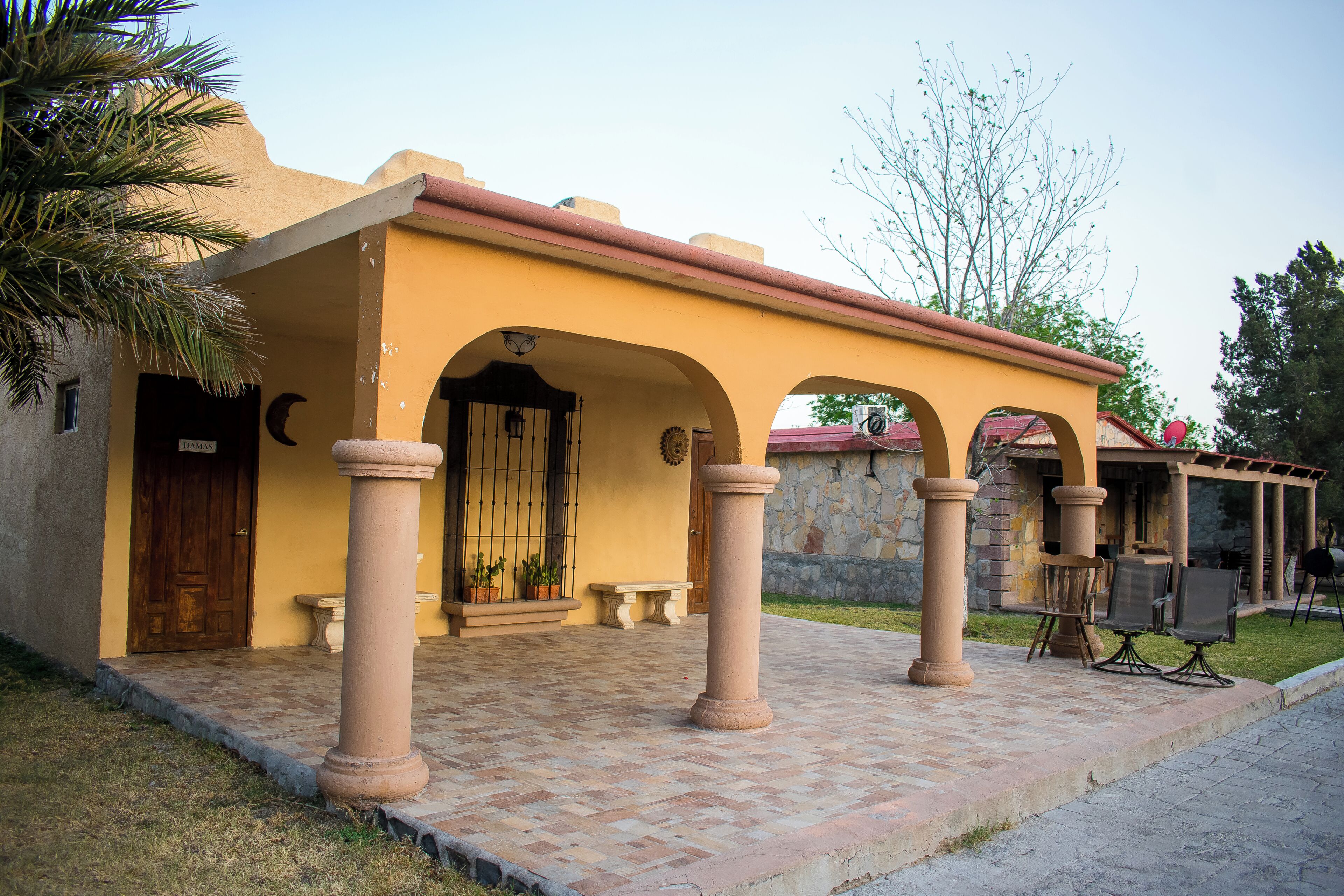 Un edificio con fachada amarilla, un acceso cubierto con columnas y un muro de piedra al fondo.