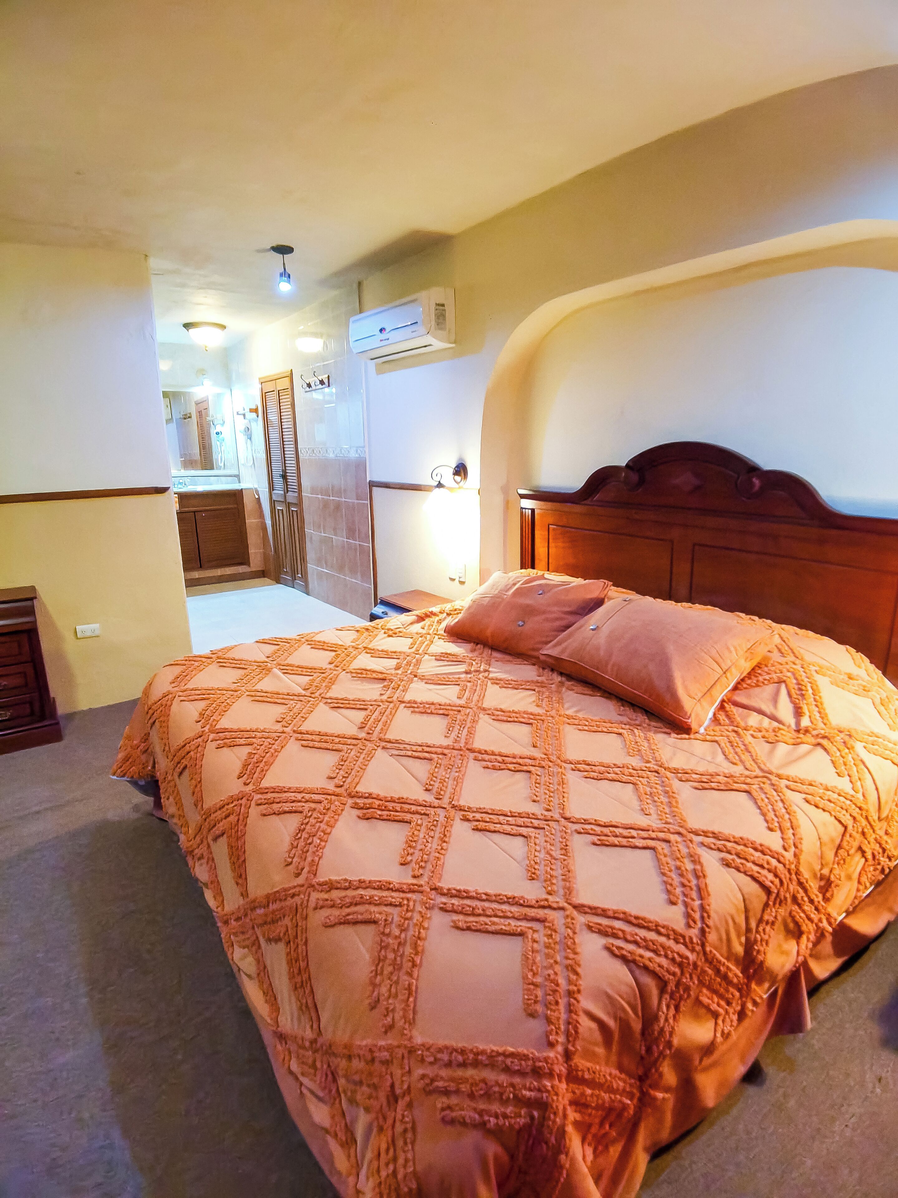 Une chambre avec un grand lit, un appareil de climatisation, une tête de lit en bois et une salle de bain visible au fond.