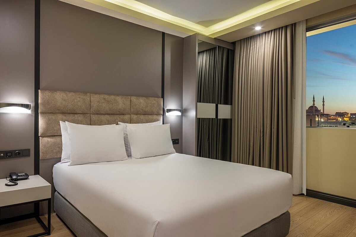 A modern hotel room with a large bed, a bedside table with a phone, and a view of the cityscape through the window.