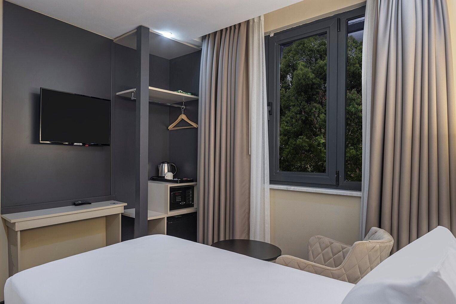 A hotel room with a bed, a desk with a flat-screen TV, a kitchenette, and a window with curtains.