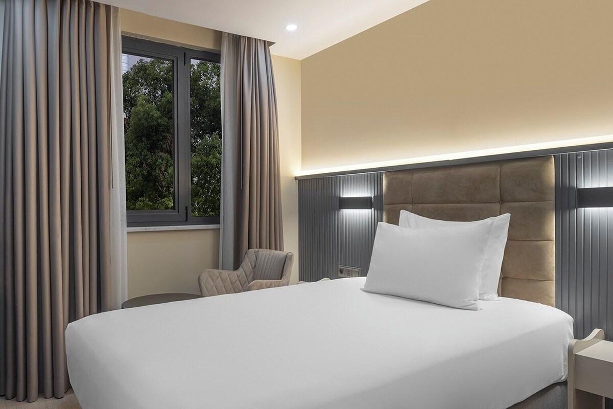 A modern hotel room with a large bed, a nightstand, a chair, and a window with curtains.