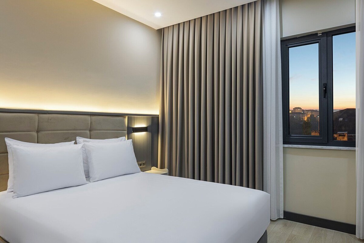 A hotel room with a large bed, a headboard, a bedside table, a lamp, a window with curtains, and a view of the cityscape.