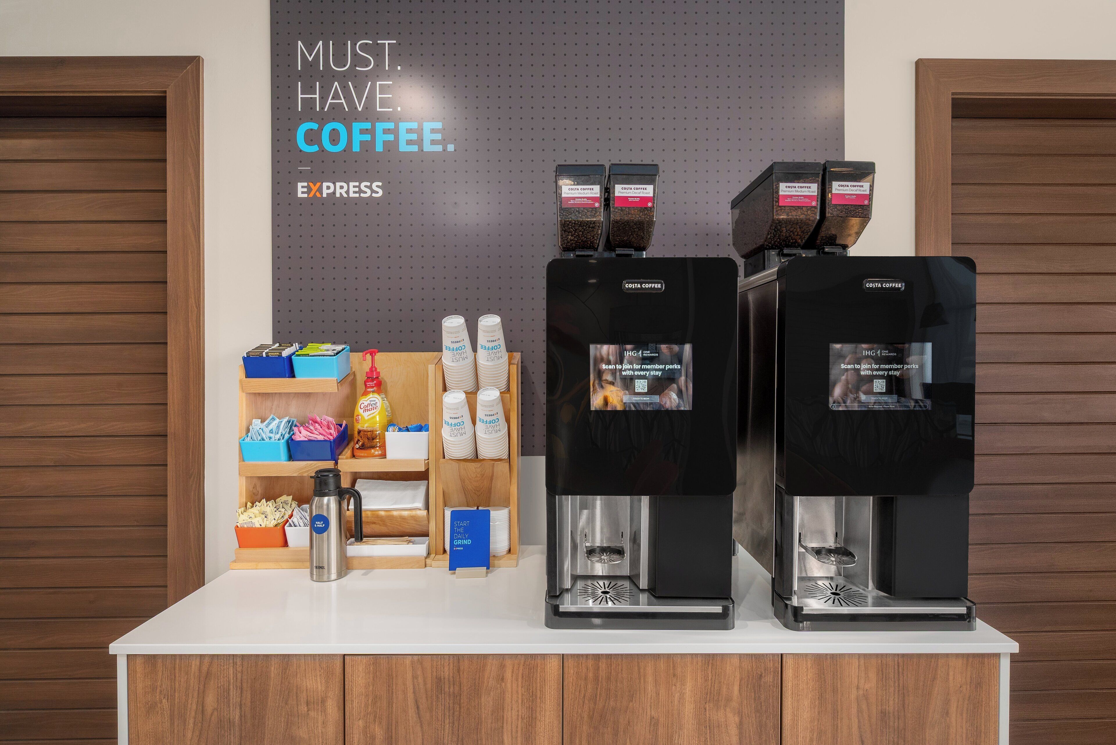 Una estación de café con varios dispensadores, un cartel que promociona la membresía y diversos productos relacionados con el café en estantes.