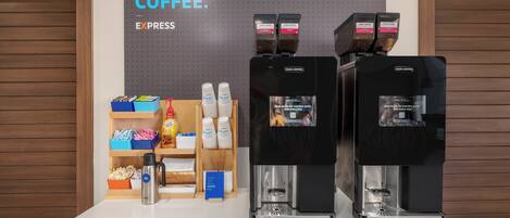Una estación de café con varios dispensadores, un cartel que promociona la membresía y diversos productos relacionados con el café en estantes.