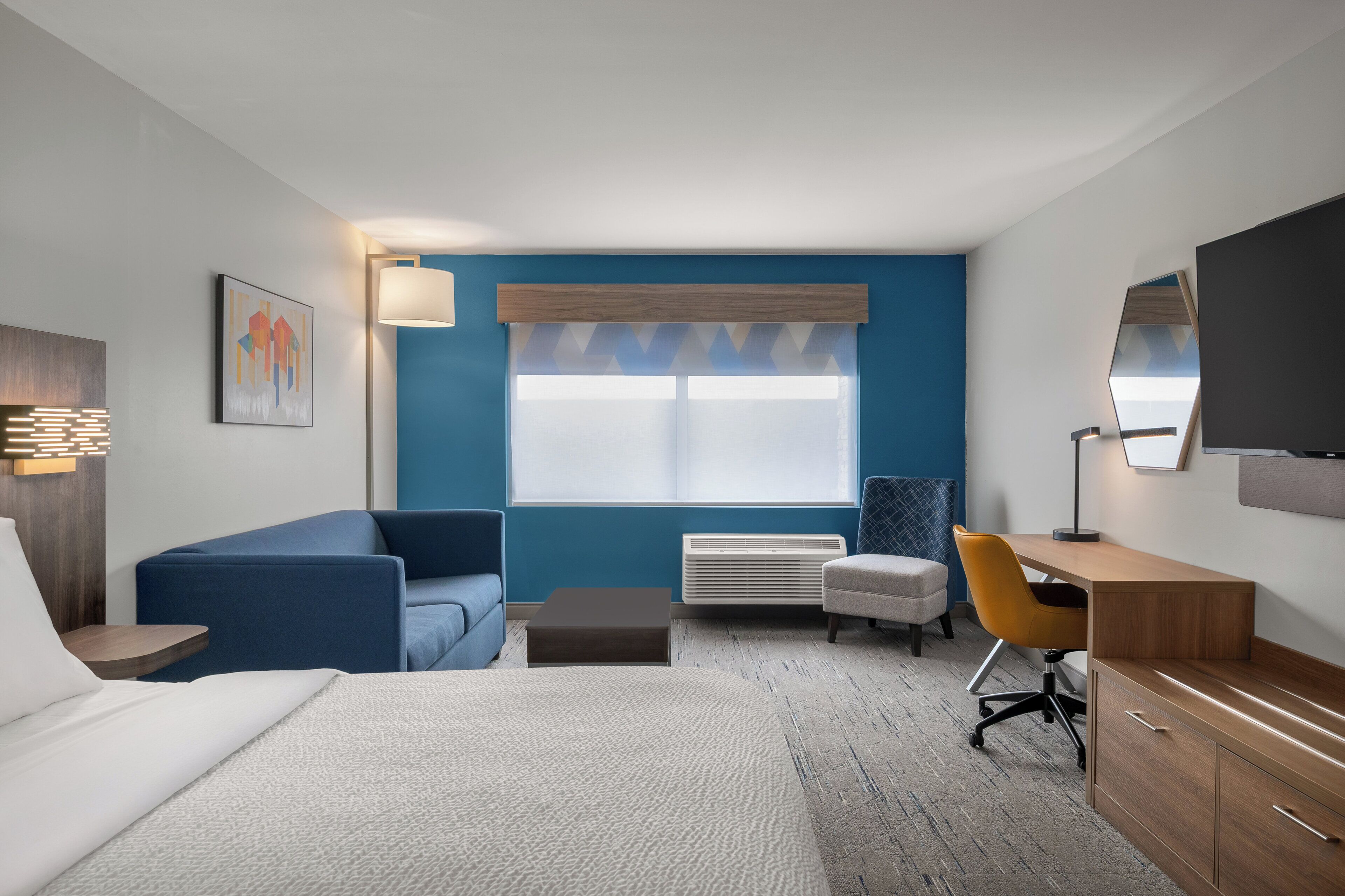 A modern hotel room with a bed, a blue sofa, a desk, a chair, a lamp, a TV, and a window with blinds.