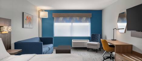 A modern hotel room with a bed, a blue sofa, a desk, a chair, a lamp, a TV, and a window with blinds.