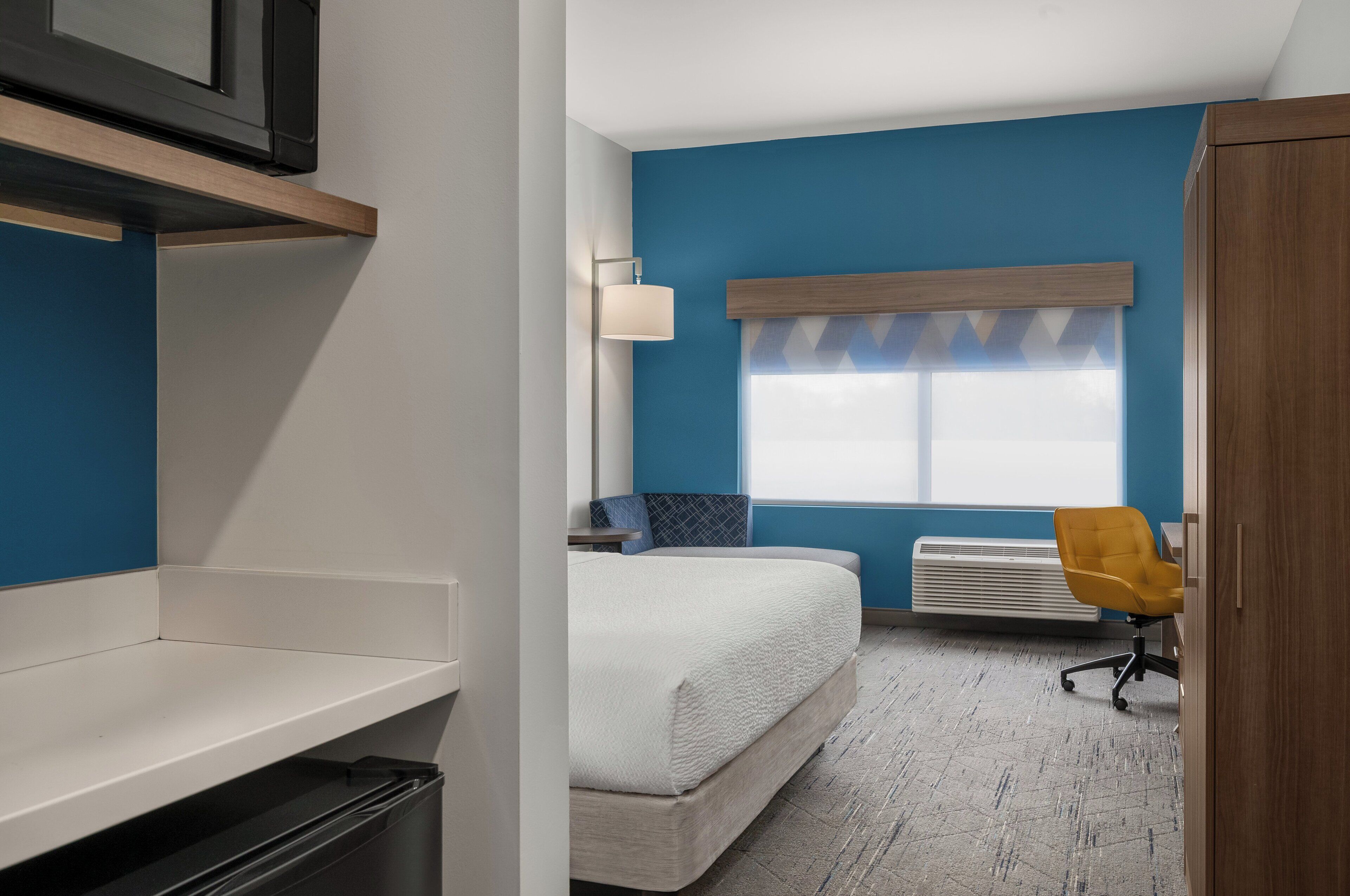 Una habitación de hotel con cama, escritorio, silla, ventana con persianas, lámpara y radiador.