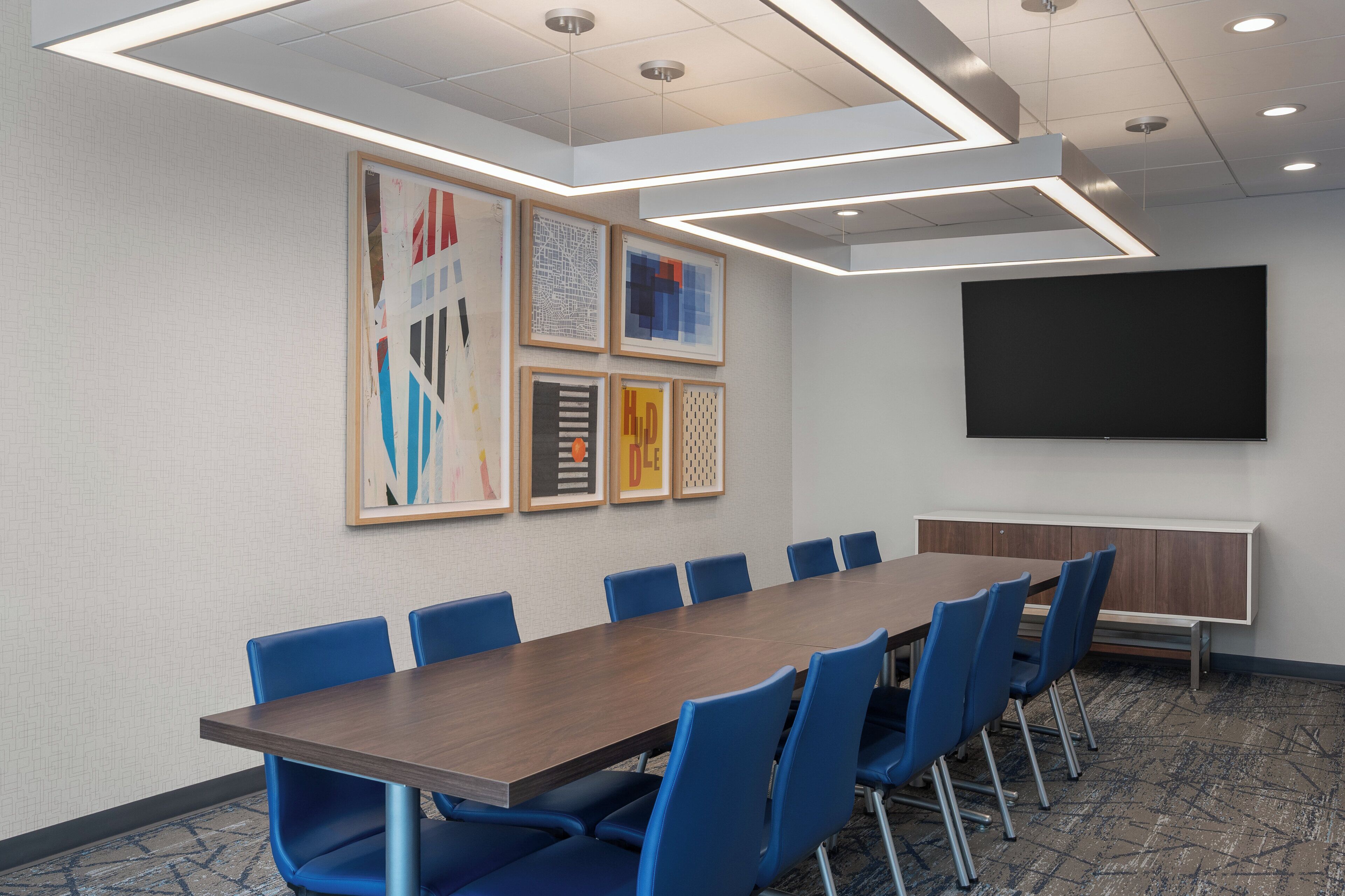 A conference room with a long table, blue chairs, framed artwork on the wall, a large screen, and recessed ceiling lighting.