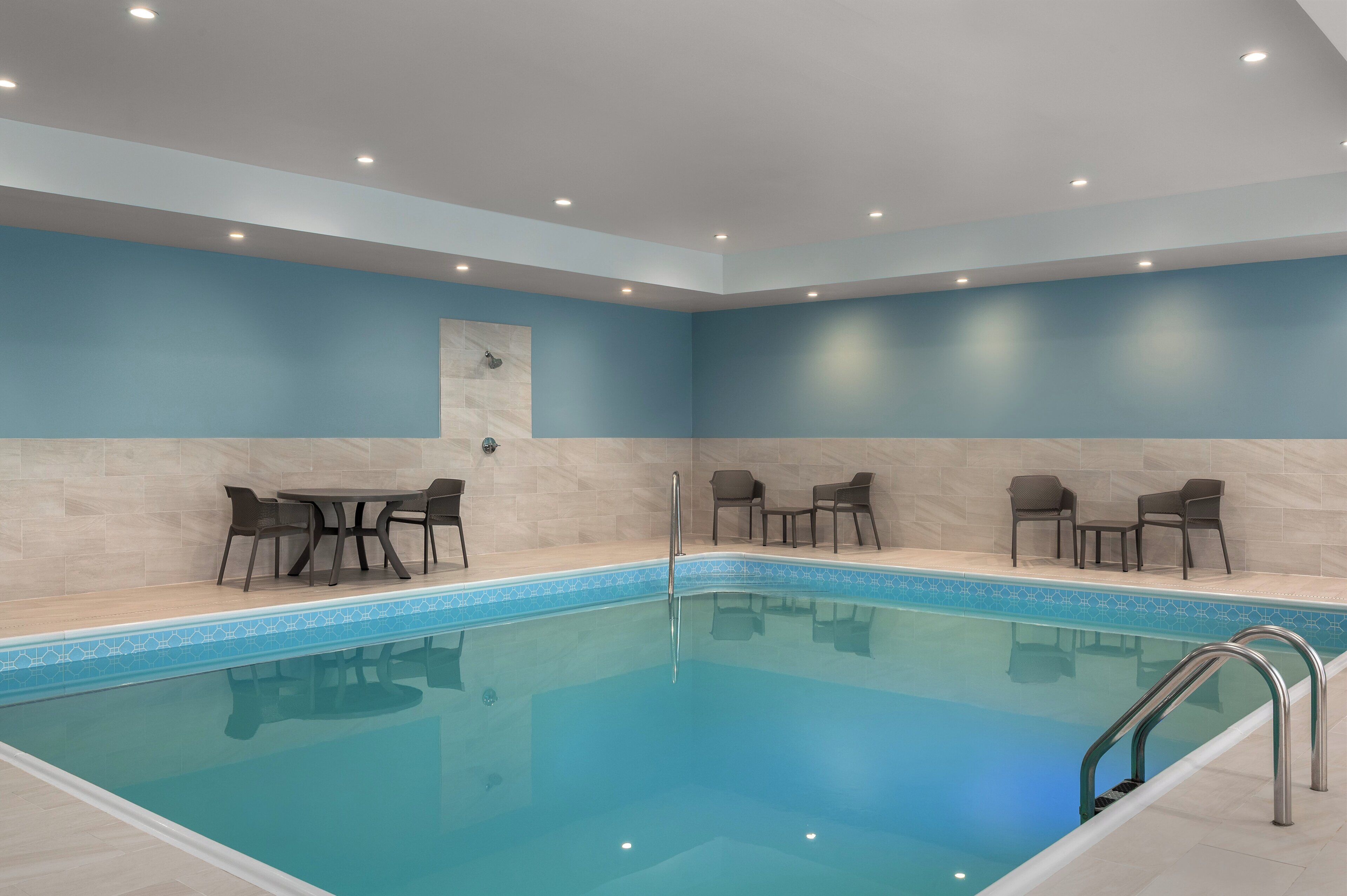 A swimming pool with a tiled edge, surrounded by chairs and tables, with a light blue wall and ceiling.