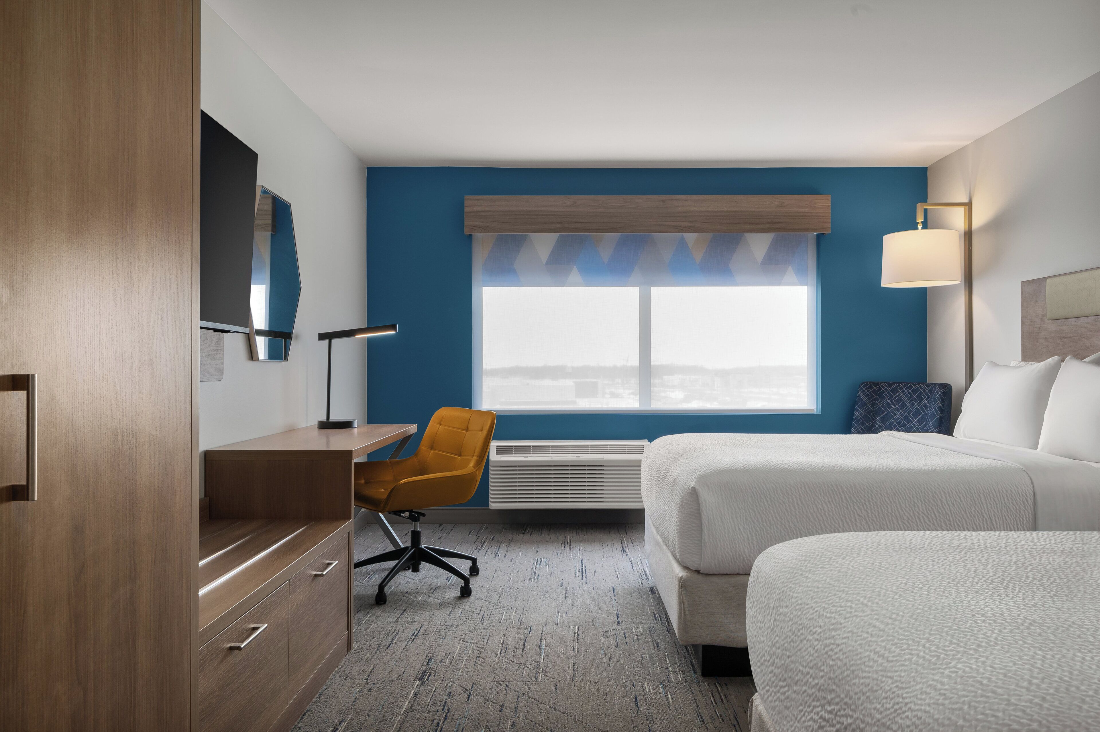 A hotel room with a desk, two beds, a chair, a large window, and a wall-mounted lamp.