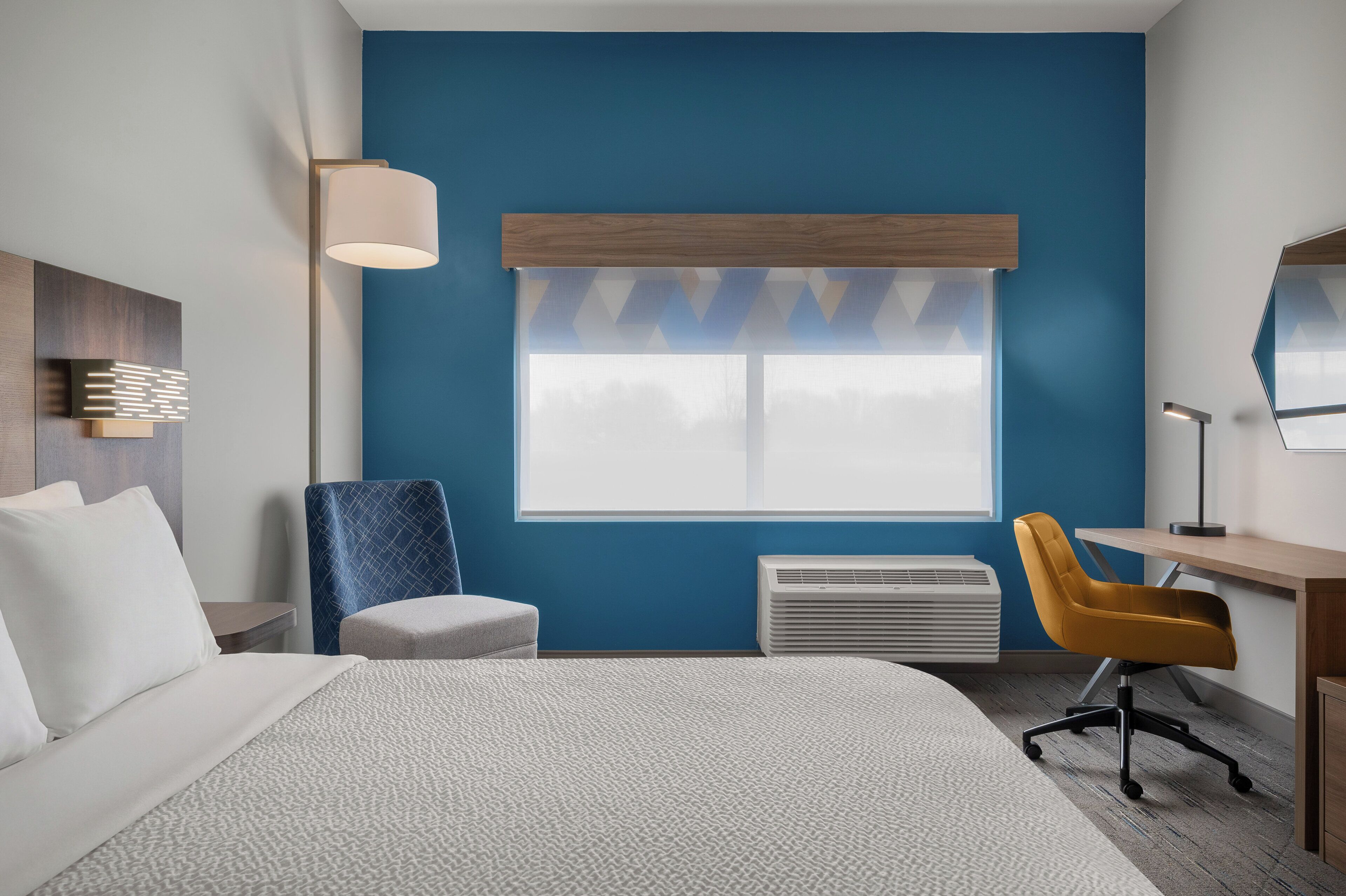 A hotel room with a bed, a desk, a chair, a standing lamp, and a window with a view.