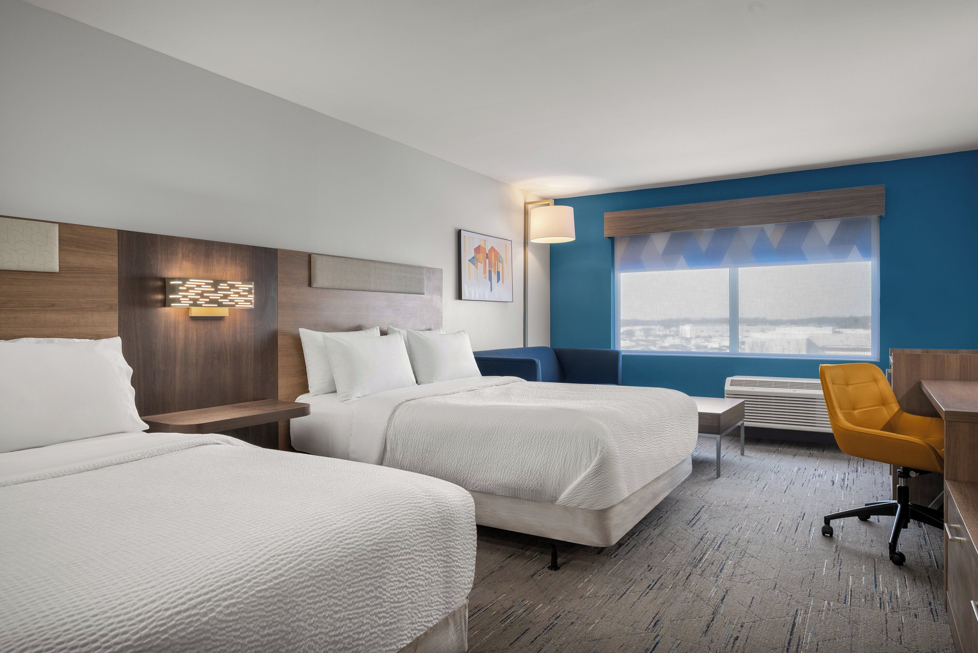 A hotel room with two beds, a desk, a chair, a window with a view, and a wall-mounted lamp.