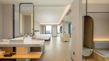 A modern hotel room with a large bed, a bedside table, a desk with a computer, a large mirror, and a view of the city through the windows.
