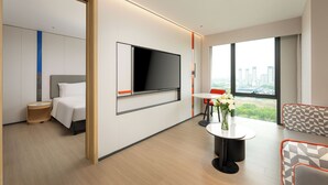 Een moderne hotelkamer met een grote televisie, een bed, een klein tafeltje met bloemen en uitzicht op de stad door grote ramen.