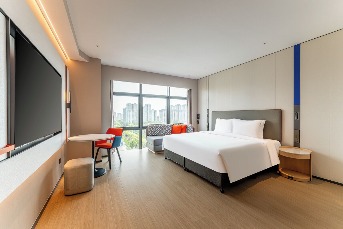Una habitación de hotel moderna con una cama grande, una zona de estar con un sofá y sillones, una mesita y un ventanal con vistas a la ciudad.